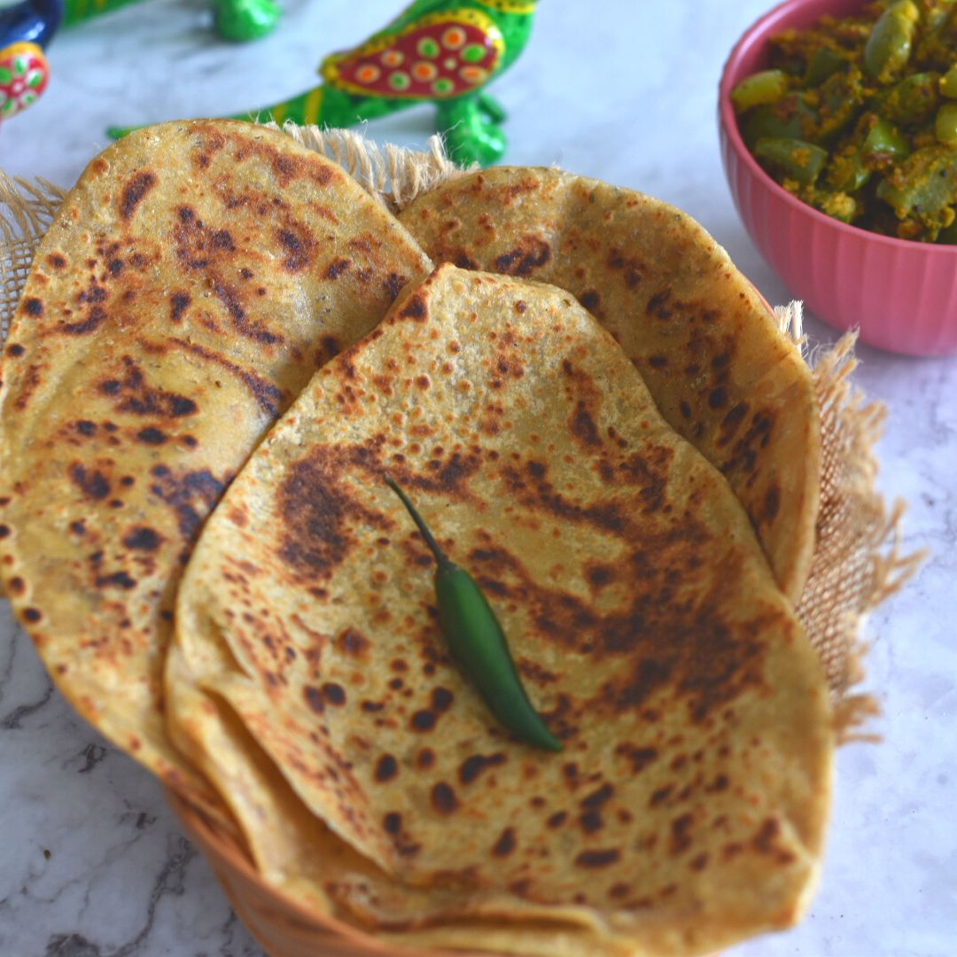 Triangular Ajwain Paratha stacked on a basket, with a green chili on it. Seen in the background is some green capsicums veg