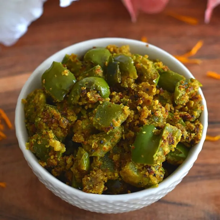 Besan Shimla Mirch or No-onion No-garlic dry capsicum veggie served in a white bowl, with some flower petals scattered and seen in the background.