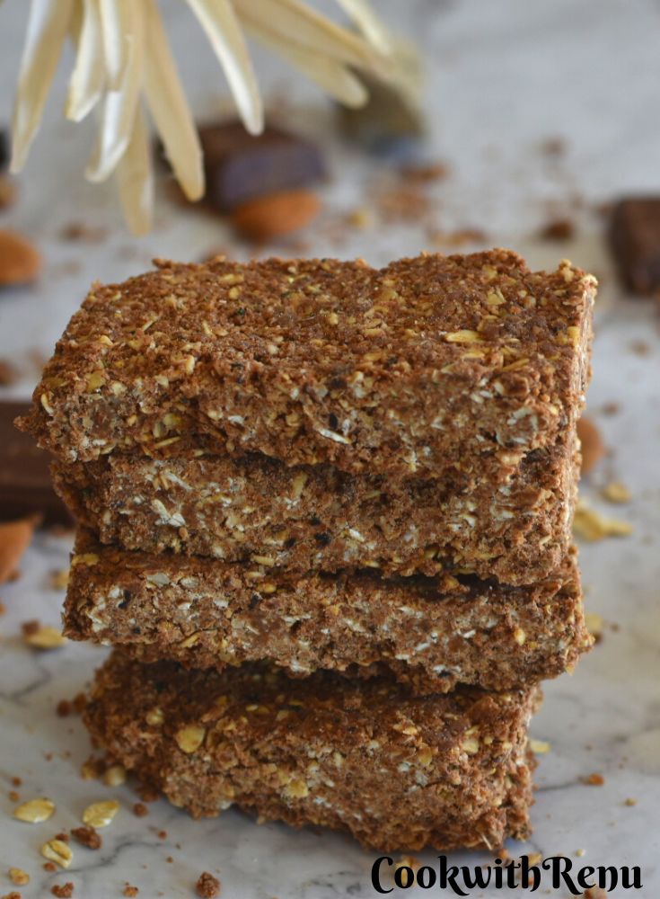 Close up view of No-Bake Oats Chocolate Protein Bar stack one above the other. Seen in the background are some chocolate, almonds and flower.