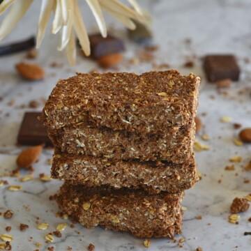 No-Bake Oats Chocolate Protein Bar stack one above the other. Seen in the background are some chocolate, almonds and flower.