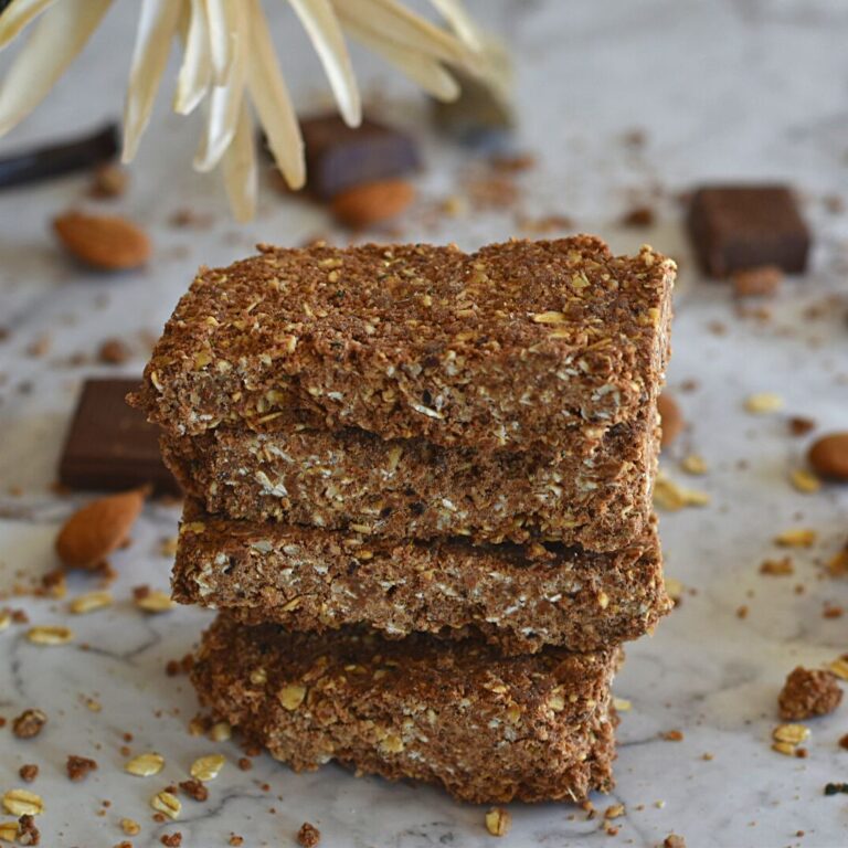 No-Bake Oats Chocolate Protein Bar stack one above the other. Seen in the background are some chocolate, almonds and flower.