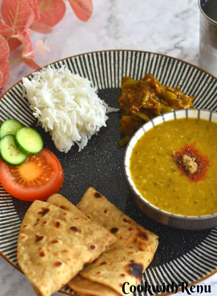Every Day Indian Thali Meal served in a black plate with Kache Tamatar Ki Sabji, moong Dal, roti, tomato, cucumber, and rice.
