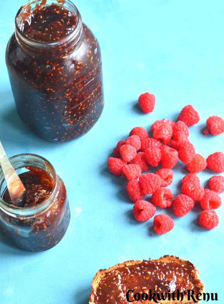Raspberry Jam spread on a slice of bread along with some raspberries scattered in background. There are 2 jam bottles filled in one small and big glass jar.