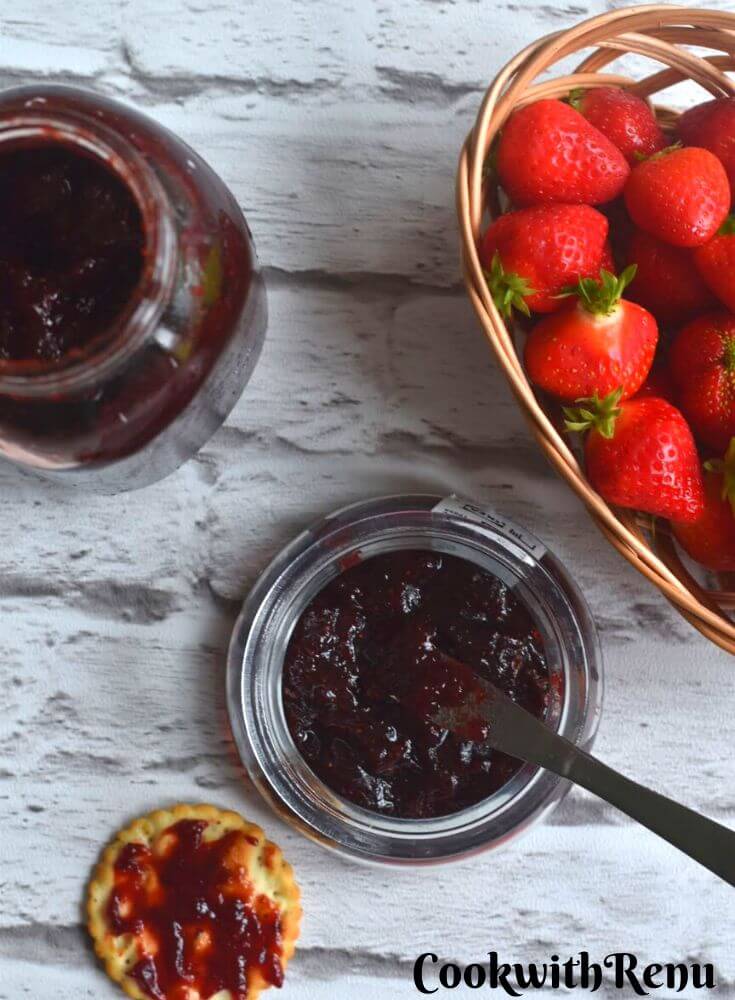 Strawberry Jam in a glass jar, with some fresh strawberries in the background in a brown punnet. In the front a cracker is spread with some jam.