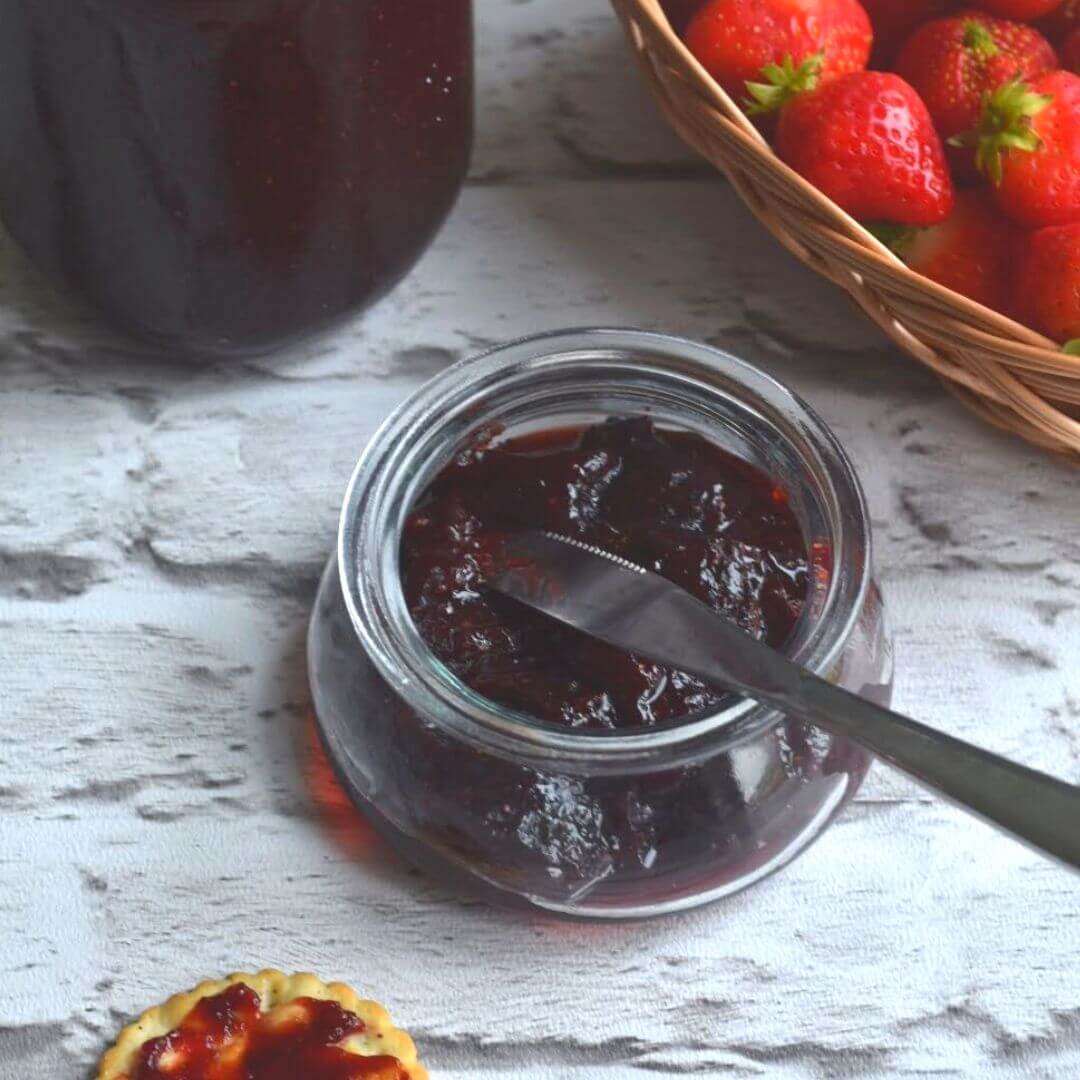 Strawberry Jam in a glass jar, with some fresh strawberries in the background.