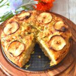 Top view of Apple and Cheddar cheese sourdough discard Focaccia on a brown board and inside the baking, with some artificial flowers behind.