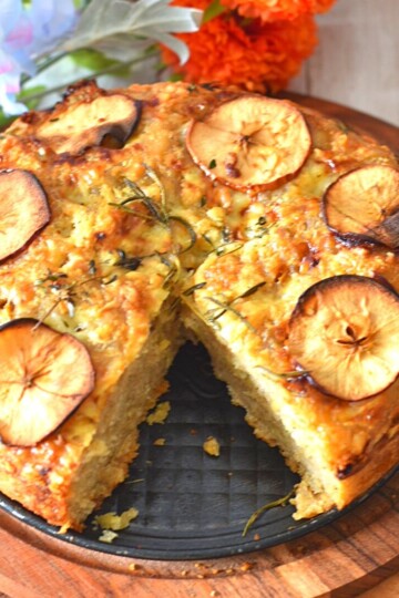 Top view of Apple and Cheddar cheese sourdough discard Focaccia on a brown board and inside the baking, with some artificial flowers behind.