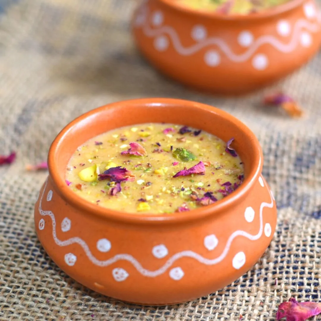 Top view of Rabri served in designer brown bowls with a garnish of rose petals and pistachio, laid on a jute cloth and rose petals scattered.