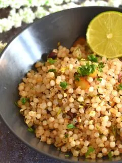 Sabudana Khichdi served in a black bowl with a garnish of lemon wedge and some flowers in the background
