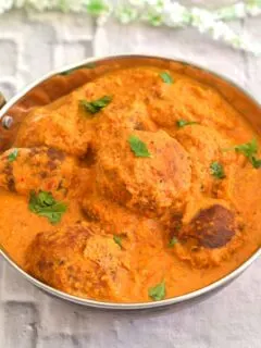 Vegetable Paneer Kofta Curry served in a steel kadai on a white board with some white flowers behind.