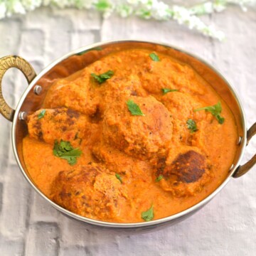 Vegetable Paneer Kofta Curry served in a steel kadai on a white board with some white flowers behind.