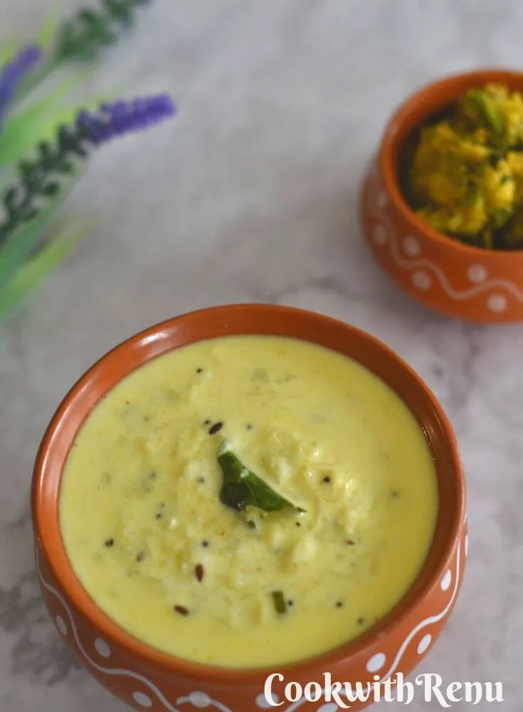 Dudhi Raita served in a brown designer bowl, with some arbi bhaji and artificial flowers seen in the background.