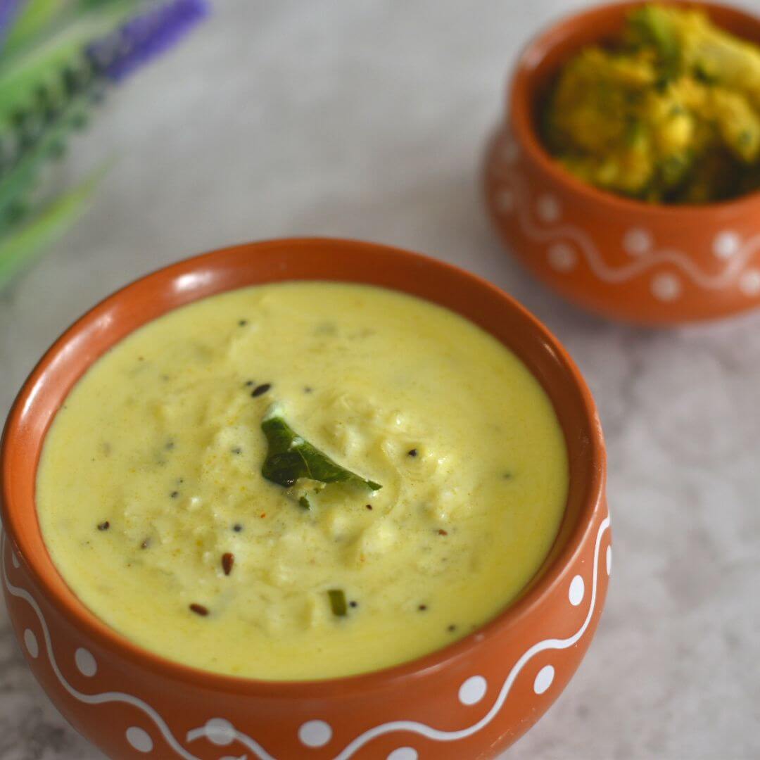 Dudhi Raita served in a brown designer bowl, with some arbi bhaji and artificial flowers seen in the background.