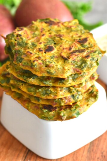Stack of Kale and Potato Fritters in a white upside-down bowl. In the background are some potatoes and kale along with a bowl of yogurt.