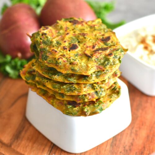 Stack of Kale and Potato Fritters in a white upside-down bowl. In the background are some potatoes and kale along with a bowl of yogurt.