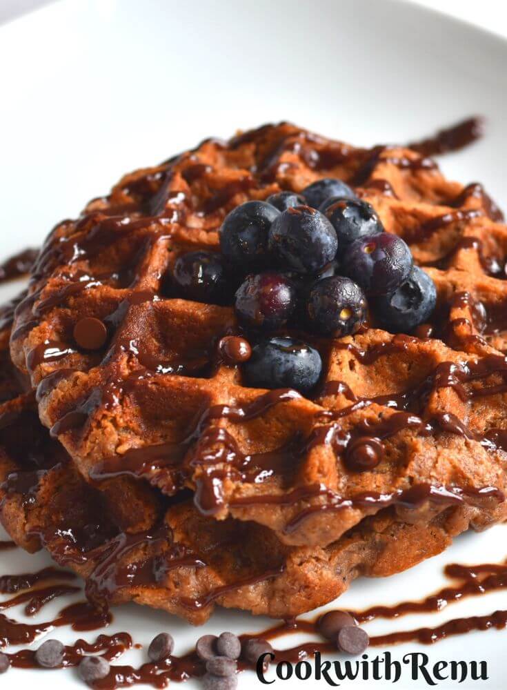 Chocolate Protein Oat Banana Waffles with a drizzle of chocolate and a garnish of chocolate chips and blueberries.