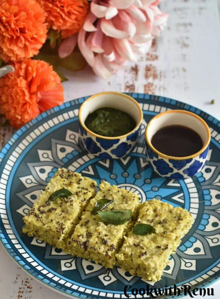 Gluten-free Millet Dhokla in a blue designer plate with some chutneys. Seen in the background are some flowers.