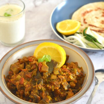 Eggplant Mushroom Stir Fry served in a white plate with grey lining. Seen in the background is a plate with lemon wedge, paratha, onion, and green chili. Also seen is a glass of buttermilk.