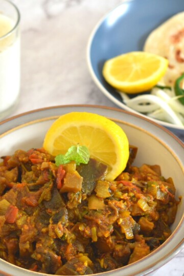 Eggplant Mushroom Stir Fry served in a white plate with grey lining. Seen in the background is a plate with lemon wedge, paratha, onion, and green chili. Also seen is a glass of buttermilk.