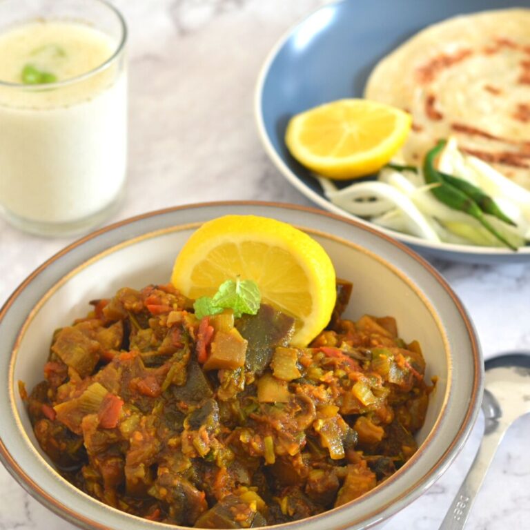 Eggplant Mushroom Stir Fry served in a white plate with grey lining. Seen in the background is a plate with lemon wedge, paratha, onion, and green chili. Also seen is a glass of buttermilk.