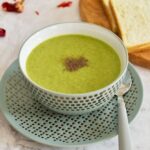 Healthy Celery and Broccoli Soup served in a green designer bowl with some bread slices in the background.