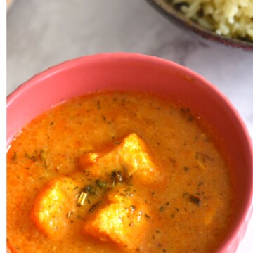 Paneer Butter Masala served in a red bowl, with Jeera rice and some salad in the background.