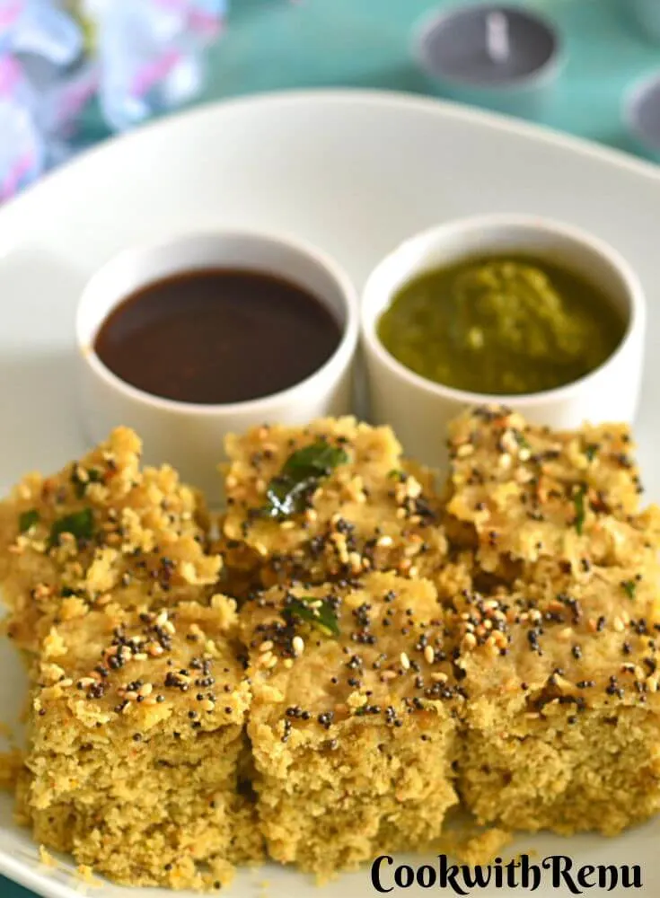 Gluten-free Millet Dhokla in a white plate with some chutneys. Seen in the background are some flowers and candles