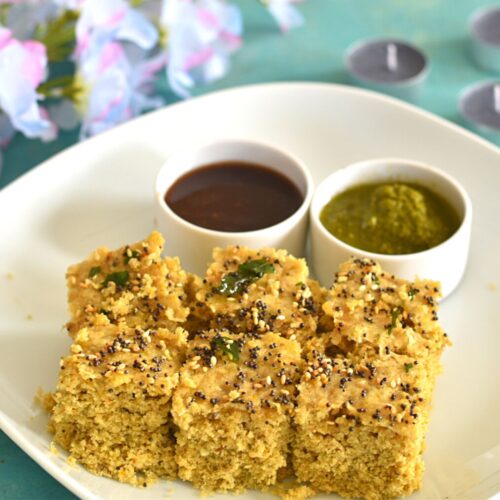 Gluten-free Millet Dhokla in a white plate with some chutneys. Seen in the background are some flowers and candles