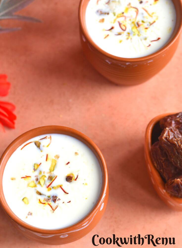 Khajoor Dudh served in 2 brown cups with some dates and flowers in the background