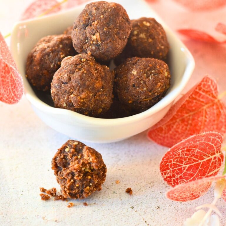 Processed Sugar-Free Ragi Ladoos in a bowl. One ladoo cut into half showing the inside texture. Some artificial flowers in the background.