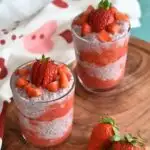 Strawberry Chia Seed Pudding presented in 2 glass, on a brown board. Seen in the background in front is some strawberries and behind is a clean towel.