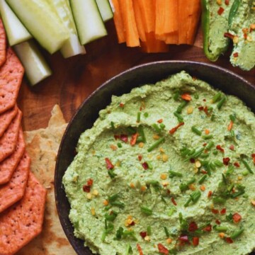 Avocado Tofu Herb Spread in a black bowl, with some crackers, veggies, and stuffed peppers seen in the background.