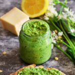 Front view of Wild Garlic Pesto in a jar, with some cheese, lemon, wild garlic and a slice of bread with pesto spread on it.