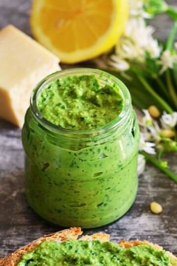 Front view of Wild Garlic Pesto in a jar, with some cheese, lemon, wild garlic and a slice of bread with pesto spread on it.