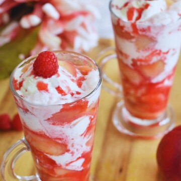 Easy Peach Melba Sundae Served in 2 glasses, with a cherry on top as garnish. Seen in the background are some flowers.
