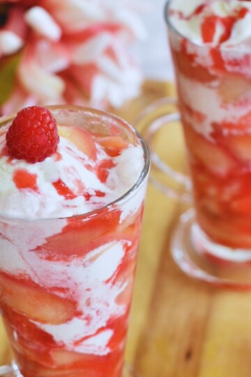 Easy Peach Melba Sundae Served in 2 glasses, with a cherry on top as garnish. Seen in the background are some flowers.
