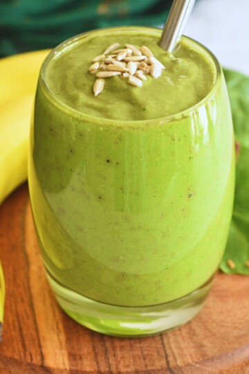 Avocado Banana Spinach Smoothie served in a glass bowl with some sunflower seeds as garnish and seen in background is banana and spinach.