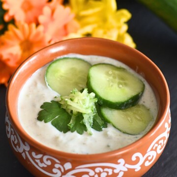 Cucumber Raita or Cucumber Yogurt sauce served in a brown bowl, with 3 small slices, and a coriander leaf and some grated cucumber as garnish. Seen in the background are some yellow flowers and cucumbers.