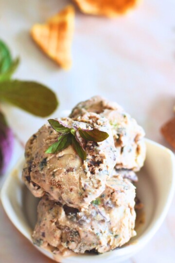 Fresh Chocolate Mint chocolate Chunk Ice Cream served in a white bowl with mint, chocolate and pieces of waffle cone behind