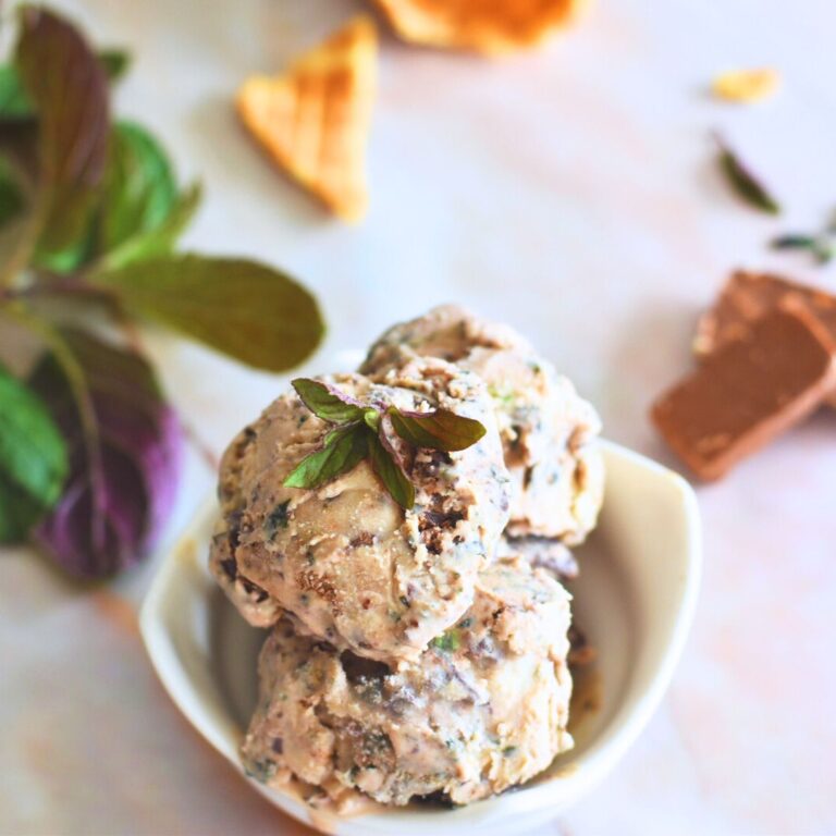 Fresh Chocolate Mint chocolate Chunk Ice Cream served in a white bowl with mint, chocolate and pieces of waffle cone behind