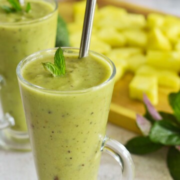Pineapple Avocado Mint Smoothie served in 2 glasses with some pineapple slices and mint in the background.