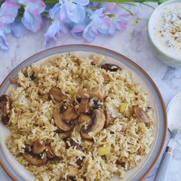 Vegan Mushroom Pulao served in a white plate with grey and brown side. Served with some raita. Some flowers seen in the background.