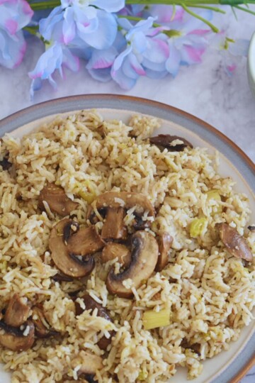 Vegan Mushroom Pulao served in a white plate with grey and brown side. Served with some raita. Some flowers seen in the background.