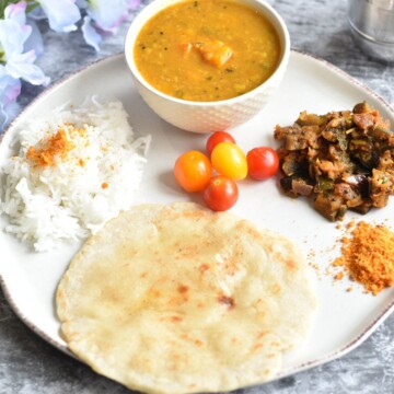 Easy Indian Vegetarian Lunch Thali served in a white plate, with sambar, eggplant stir fry, red chili chutney, rice bhakri, and rice. Seen in the background are some artificial flowers.