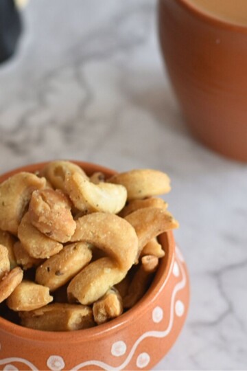 Close up view of Namkeen Kaju Matri or Namak Para served in a brown designer bowl with a cup of tea and teapot seen in the background.