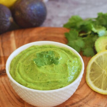 Spicy Avocado Coriander chutney served in a white bowl, with some lemon slices, coriander, avocado in the background.