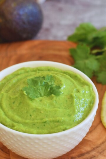 Spicy Avocado Coriander chutney served in a white bowl, with some lemon slices, coriander, avocado in the background.