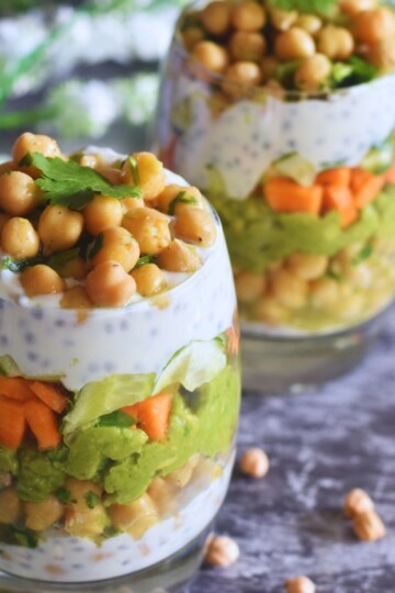 Chickpeas Avocado Lunch Parfait served in 2 glasses. Seen in the background are some chickpeas and some flowers.
