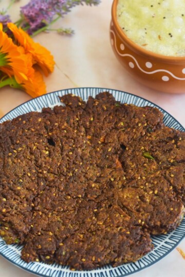 Ragi Cucumber Thalipeeth served on a blue plate along with sabudana kadhi. Seen in the background are some flowers.