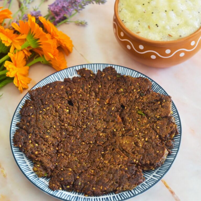 Ragi Cucumber Thalipeeth served on a blue plate along with sabudana kadhi. Seen in the background are some flowers.
